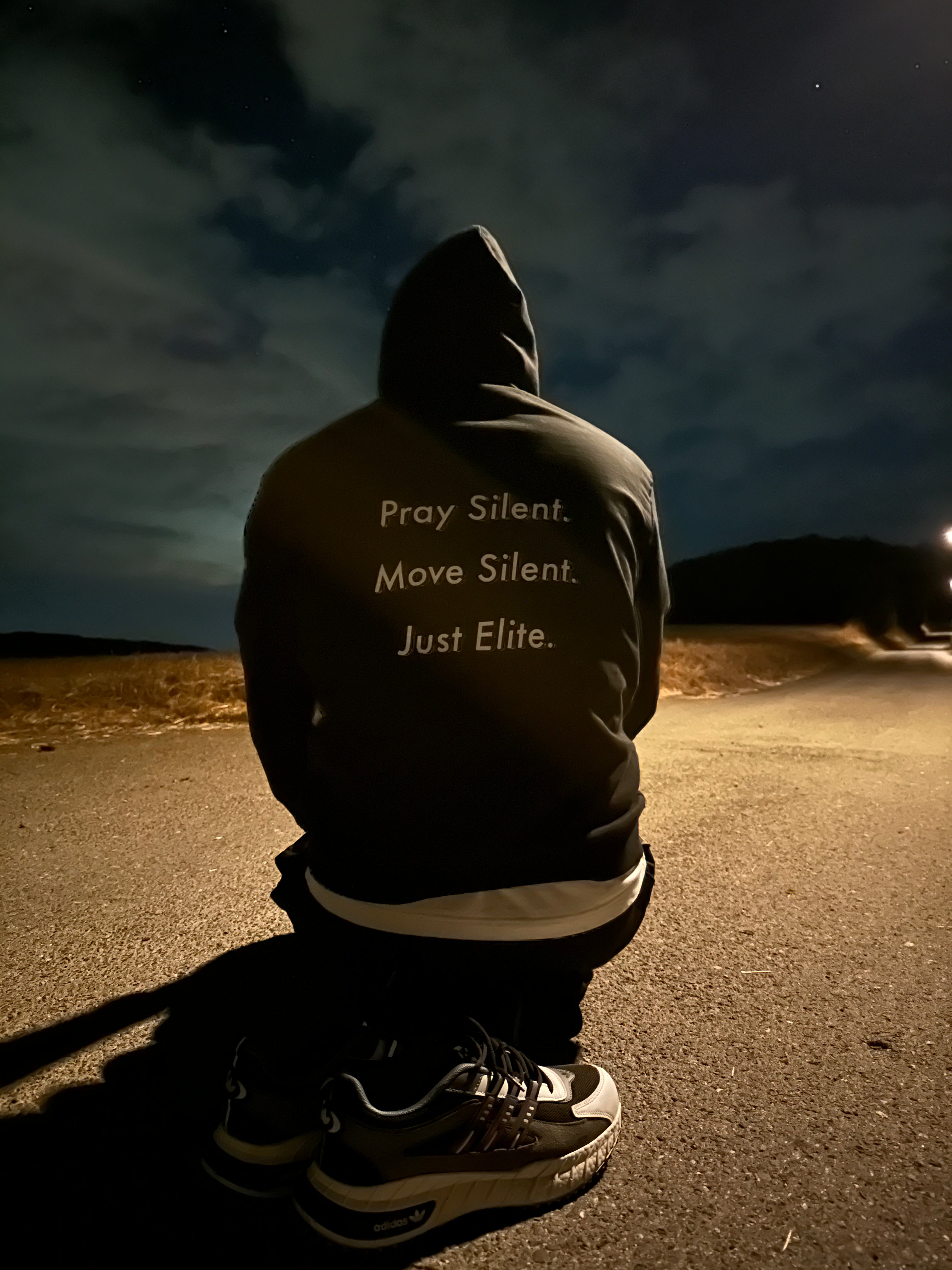Person kneeling on an empty road at night wearing the Dunya Lions hoodie with the back print: Pray Silent. Move Silent. Just Elite. A stormy sky and distant hills set a powerful, silent tone.
