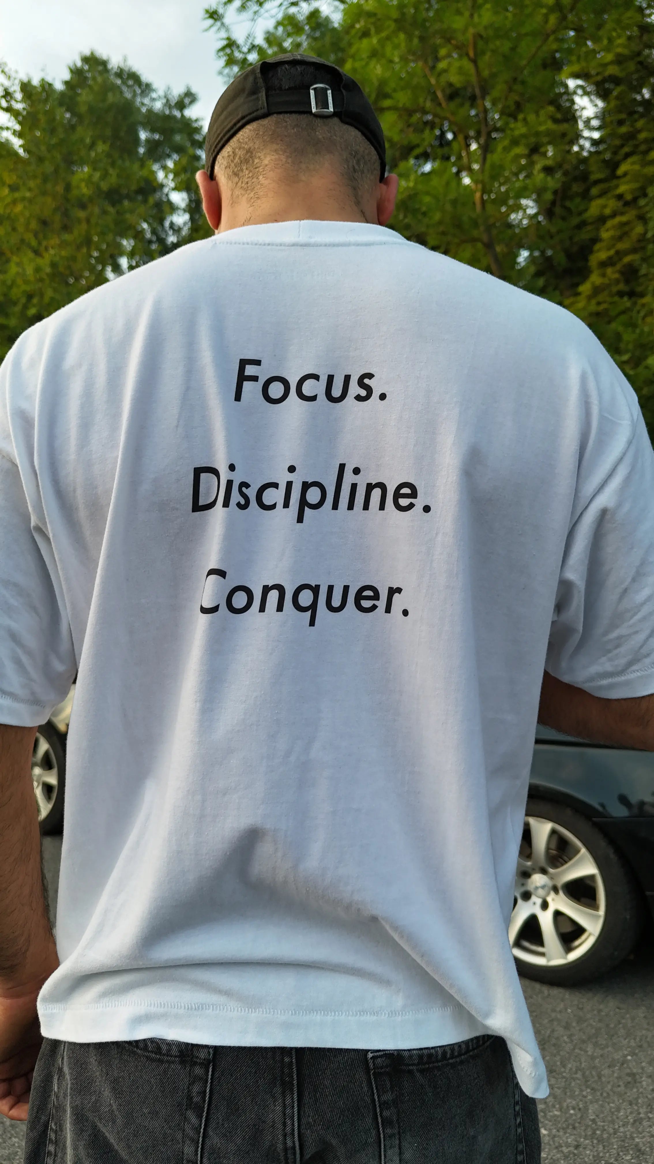 Man standing with his back to the camera, wearing the Dunya Lions white T-shirt with the message: Focus. Discipline. Conquer. A powerful street presence in daylight.