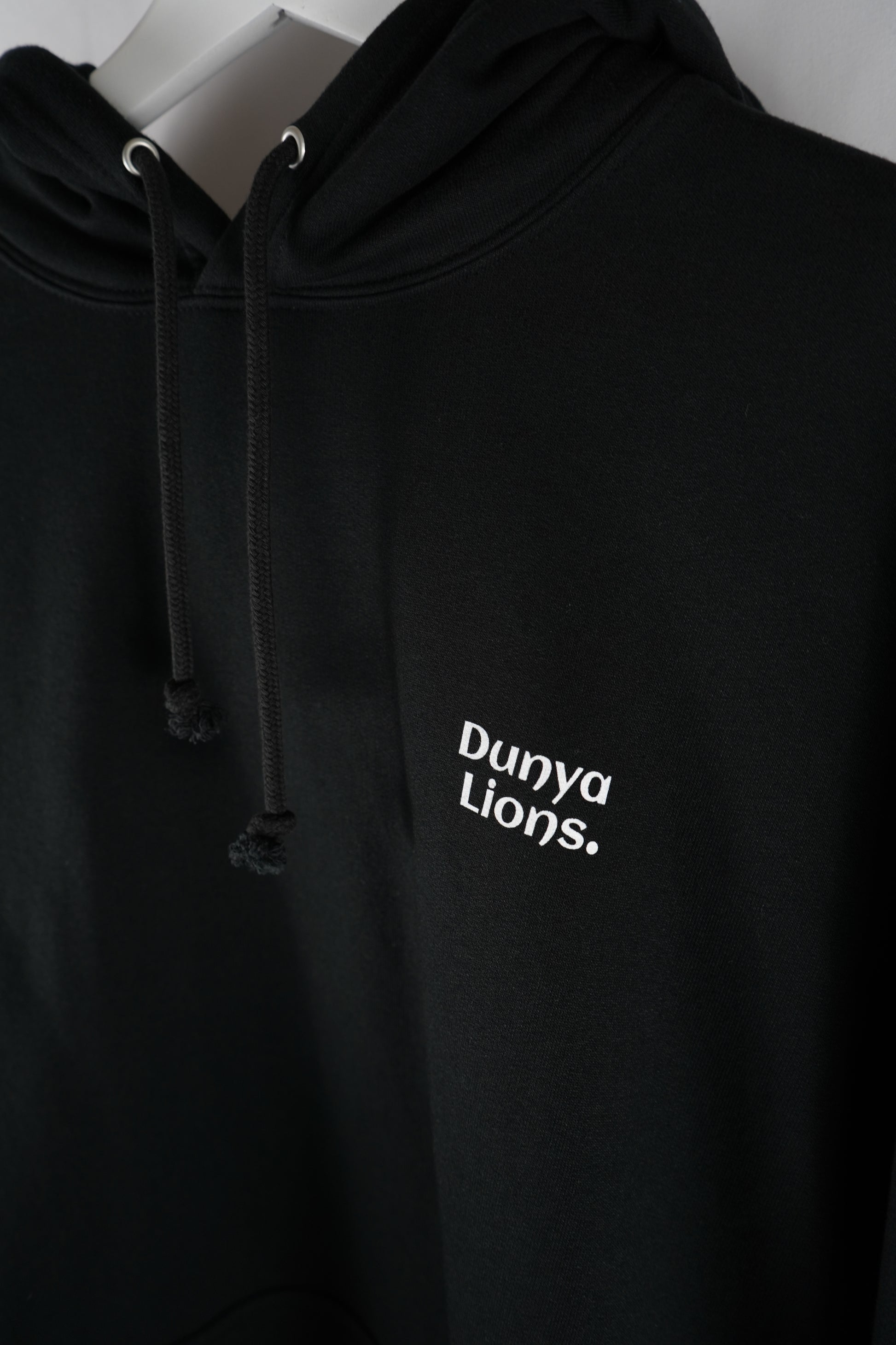 Close-up of black Dunya Lions hoodie showing the white logo on the left chest – hoodie strings visible, shot on hanger in soft studio light.