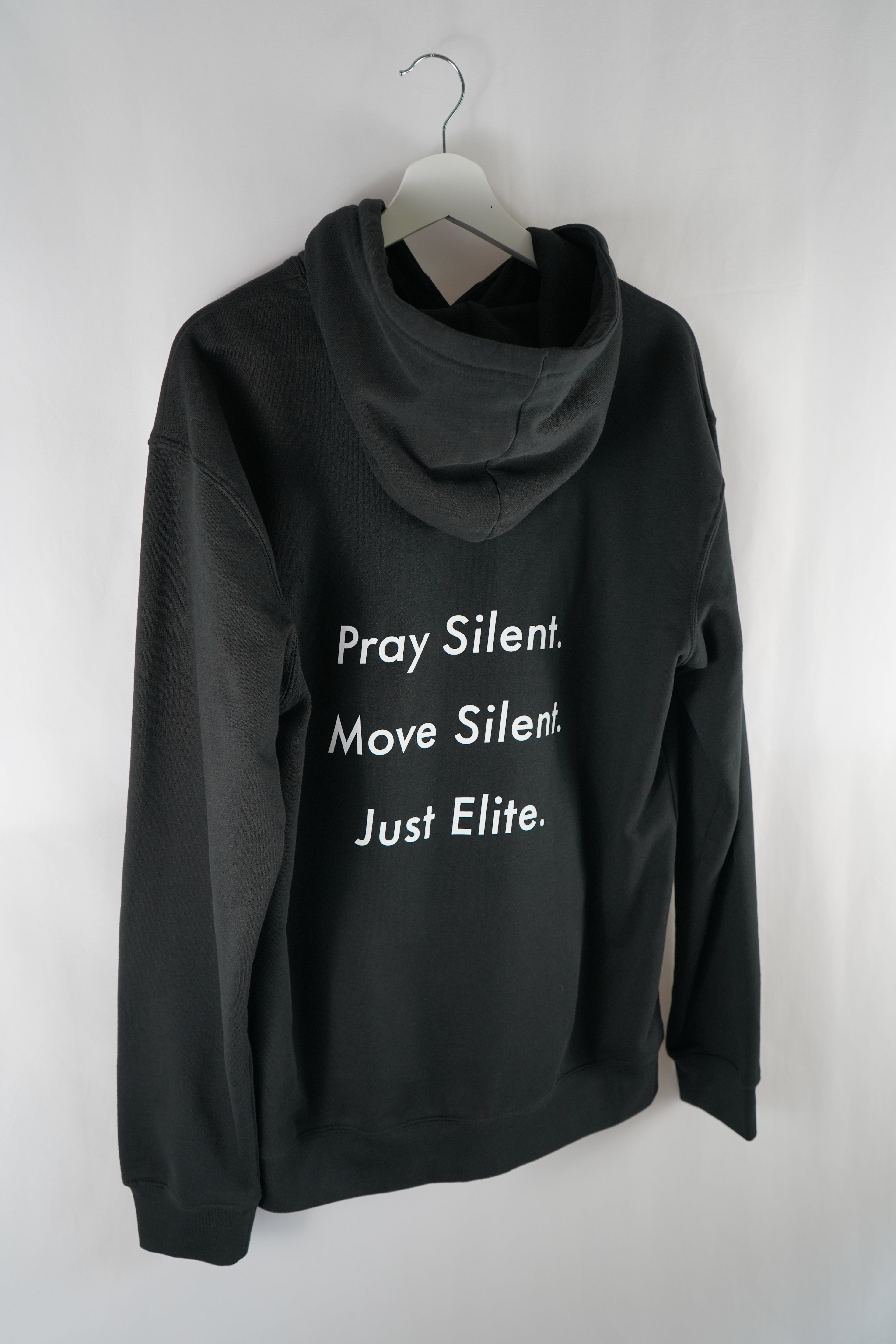 Back view of black Dunya Lions hoodie with bold white text: "Pray Silent. Move Silent. Just Elite." – hanging on a white hanger, studio-lit.
