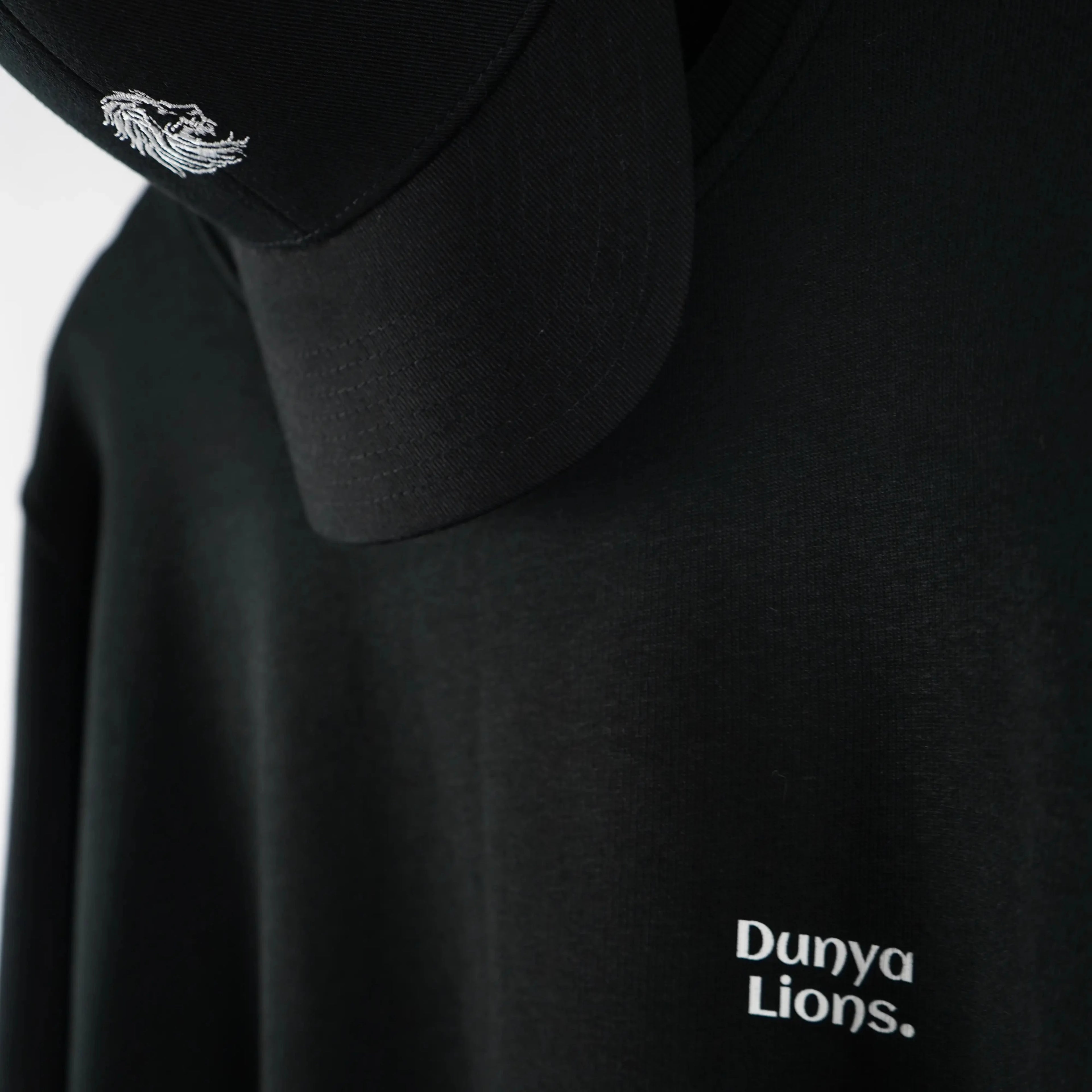 Nahaufnahme des schwarzen Dunya-Lions-Sweaters mit weißem Brustlogo und Cap mit gesticktem Löwenkopf – Symbol für stille Dominanz und klare Präsenz.