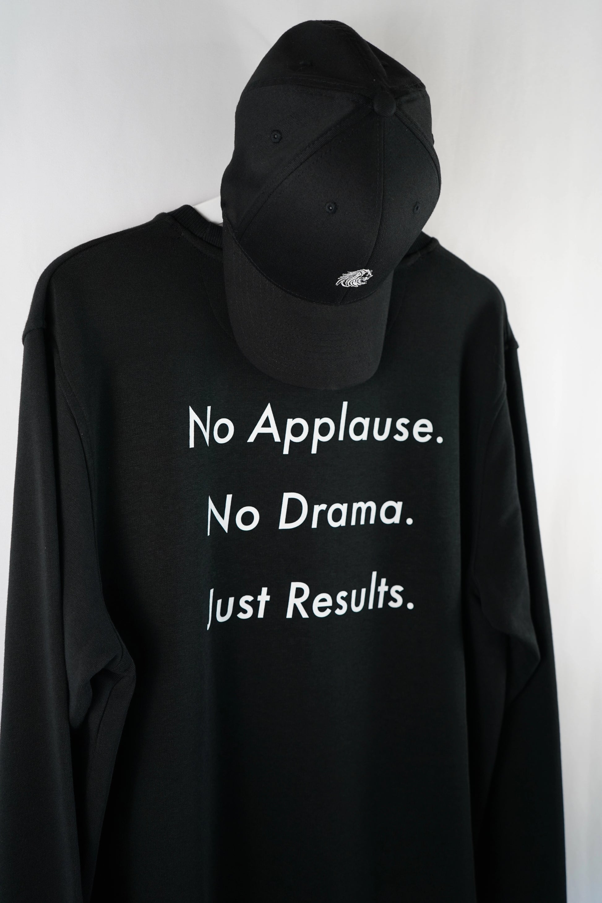 Schwarzer Dunya-Lions-Sweater mit dem Rückenprint „No Applause. No Drama. Just Results.“ – präsentiert mit schwarzer Cap, Fokus auf Message, Ruhe und Stärke.
