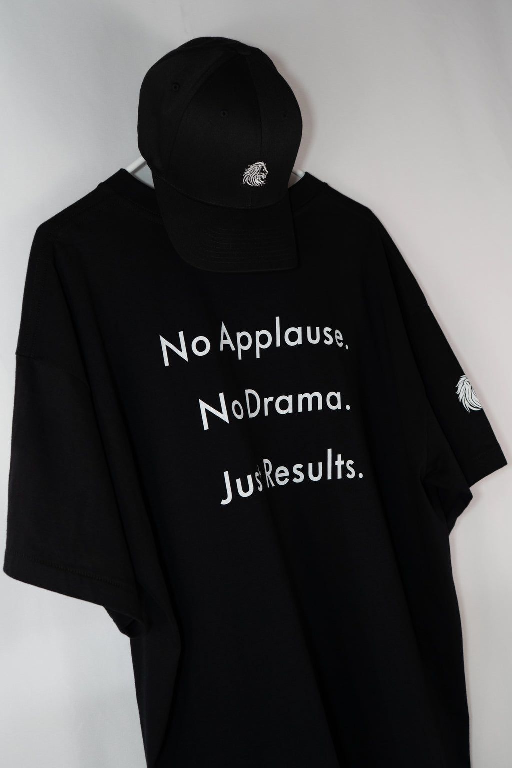 Schwarzes Oversize-T-Shirt mit weißem Statement-Print ‚No Applause. No Drama. Just Results.‘, kombiniert mit schwarzer Cap – minimalistisch aufgehängt vor hellem Hintergrund. Symbol für Ruhe, Stärke und Fokus.