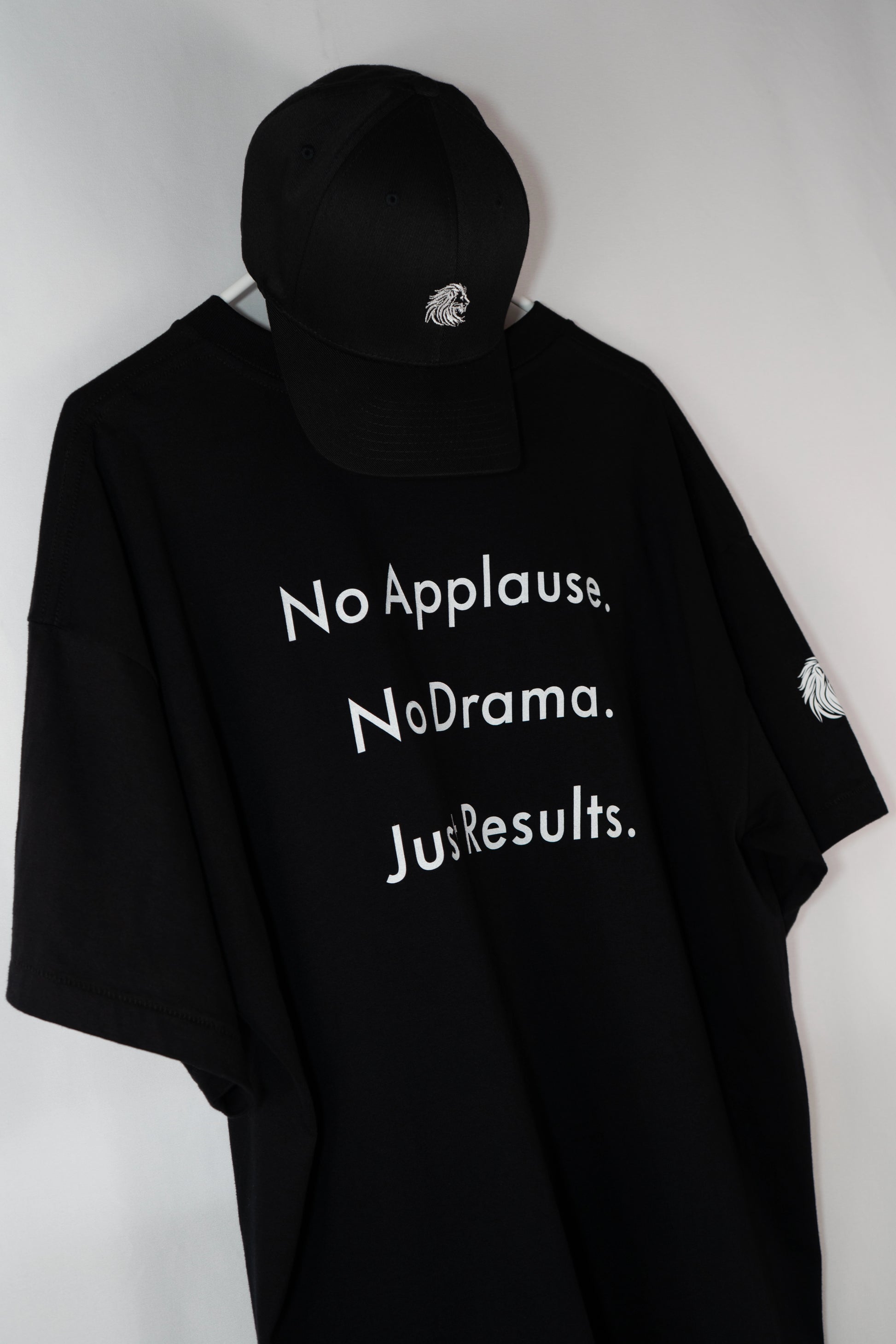 Schwarzes Oversize-T-Shirt mit weißem Statement-Print ‚No Applause. No Drama. Just Results.‘, kombiniert mit schwarzer Cap – minimalistisch aufgehängt vor hellem Hintergrund. Symbol für Ruhe, Stärke und Fokus.