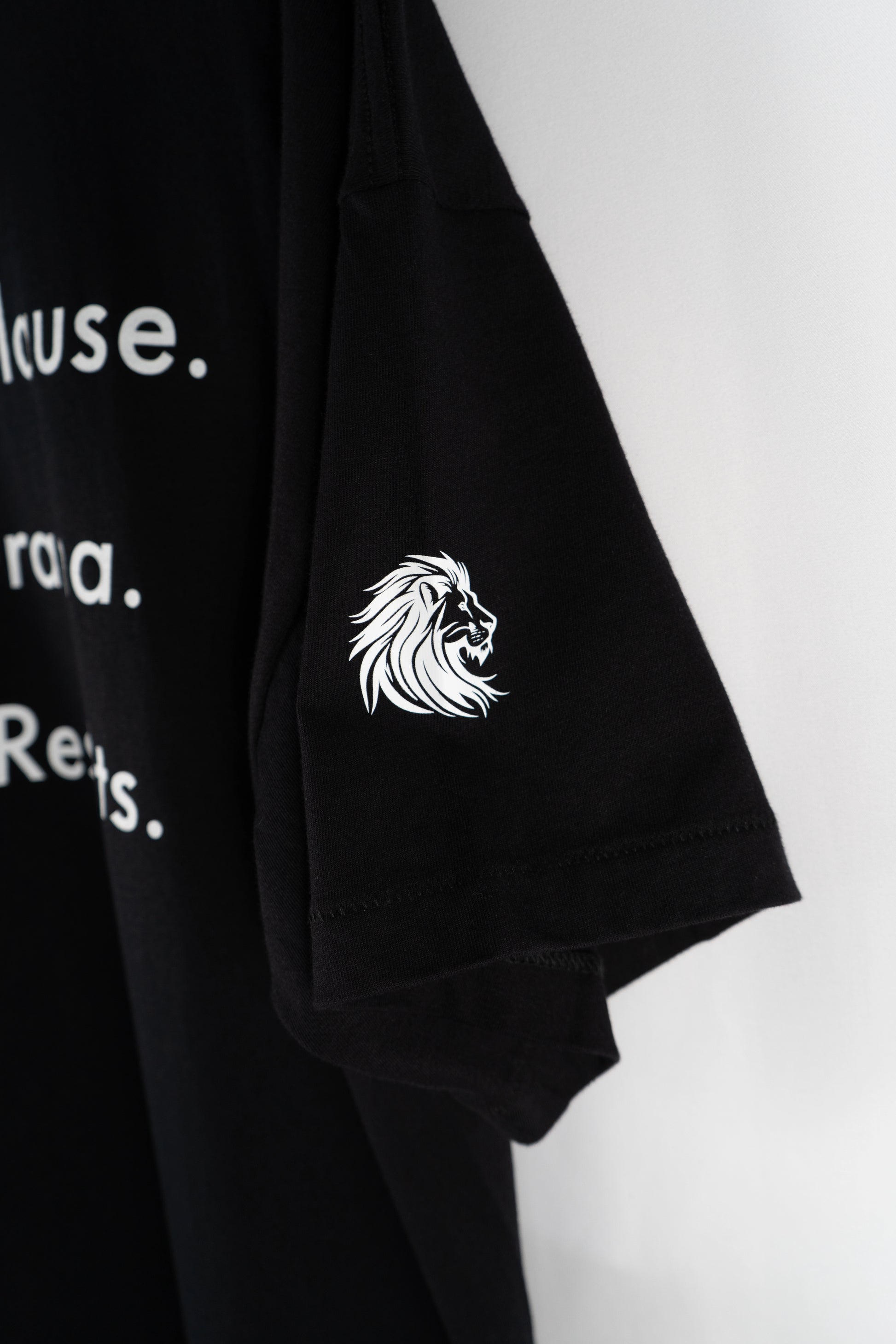 Schwarzes Oversized-T-Shirt von Dunya Lions mit weißem Löwenlogo auf dem Ärmel und dem Statement ‚No Applause. No Drama. Just Results.‘ – aufgehängt vor hellem Hintergrund. Symbol für Fokus, Disziplin und Elite-Ruhe.